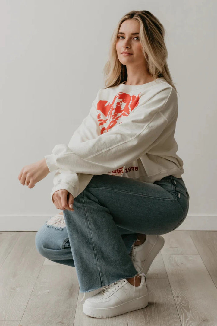 A woman in a white Rolling Stones NYC sweatshirt and jeans looking relaxed and stylish. People of Leisure Sweatshirt